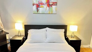 A neatly made bed with white linens and two bedside tables with lamps, a modern abstract painting on the wall, and a wooden floor. - Central 2BR | Near Metro & Parks (Montréal)