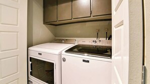 A compact laundry room with a front-loading washer and dryer, a small countertop with a sink, and a cabinet above the appliances. - Central 2BR | Near Metro & Parks (Montréal)