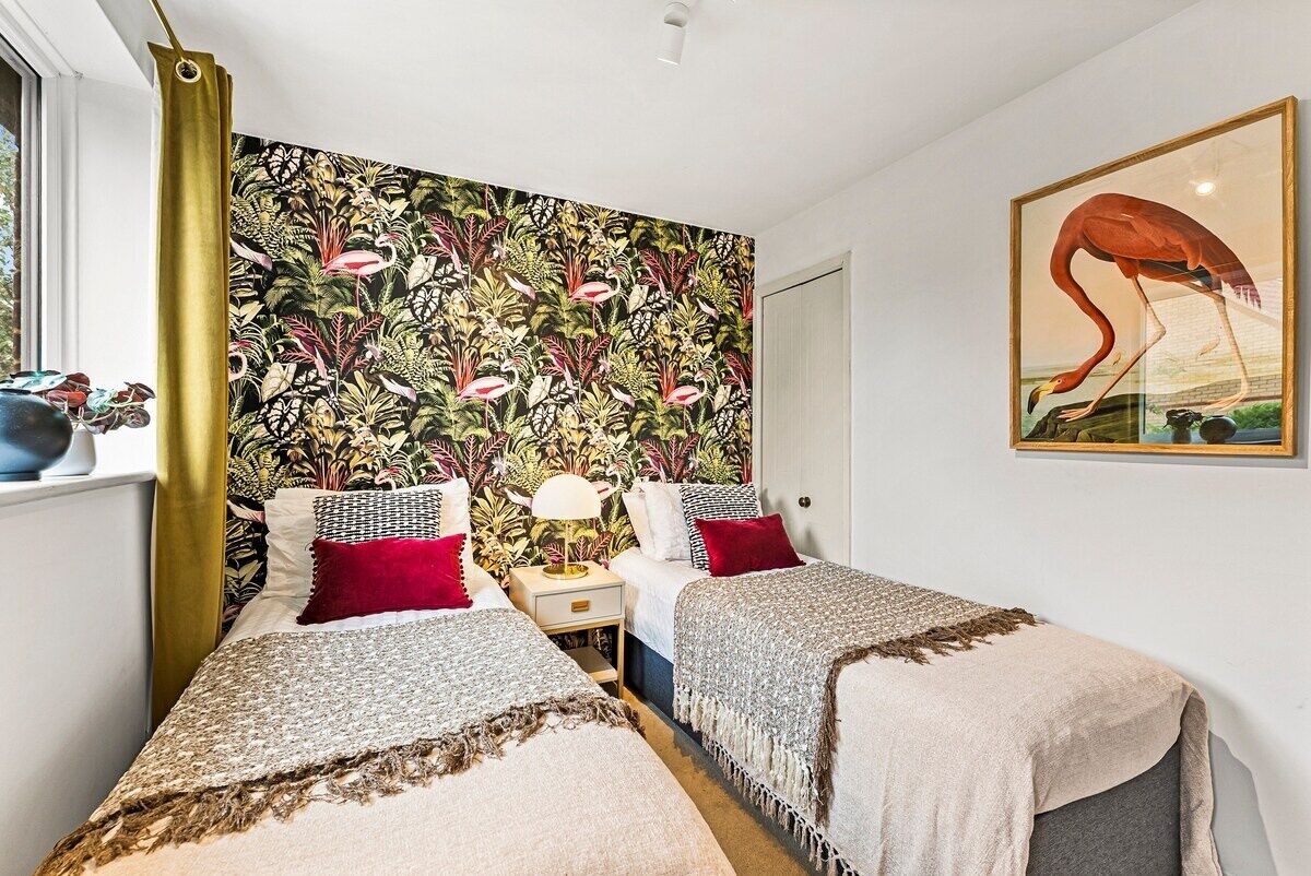 A bedroom with two beds, a tropical wallpaper, a framed painting, and a window with a curtain.