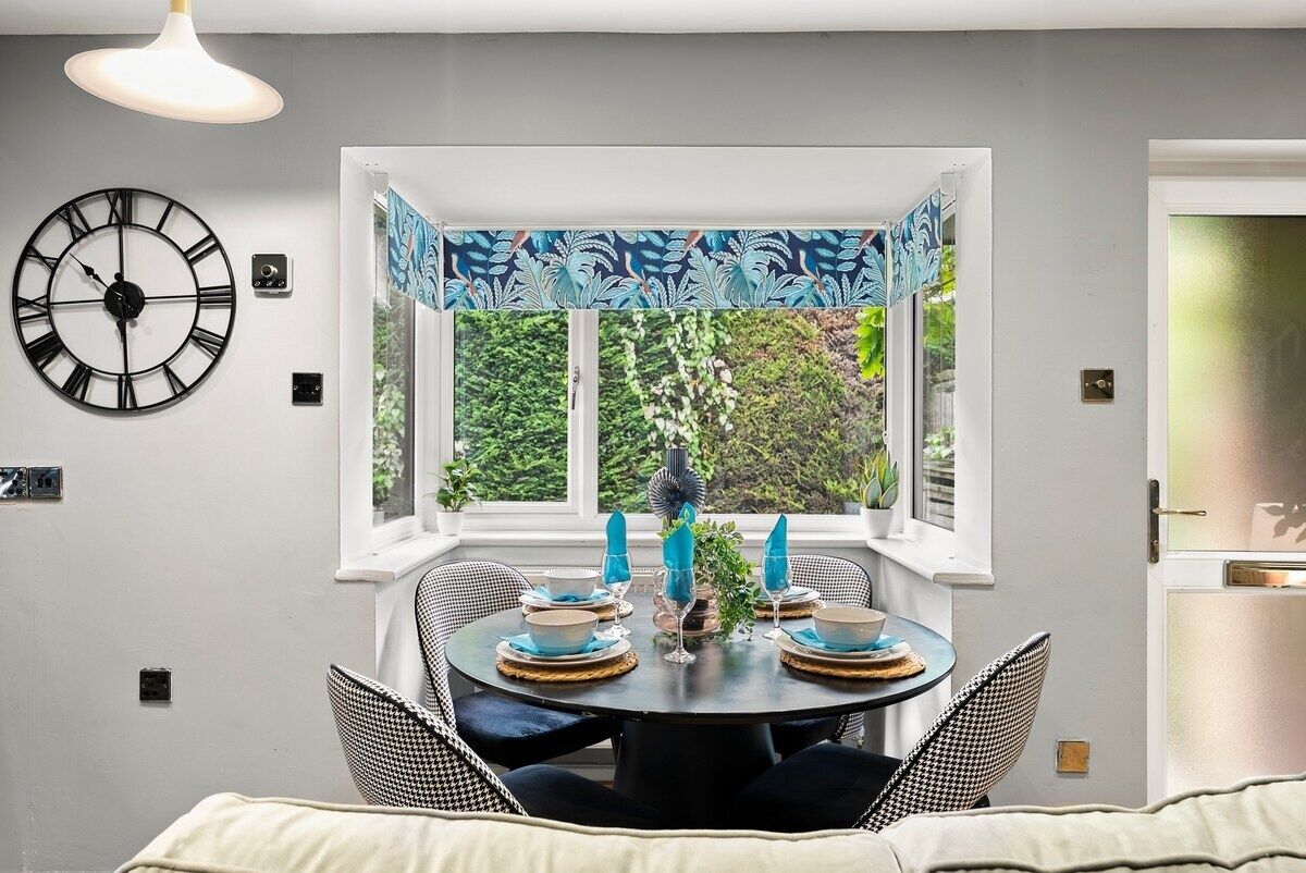 Un coin repas avec une table ronde dressée pour quatre personnes, une horloge au mur et une fenêtre donnant sur un paysage verdoyant.