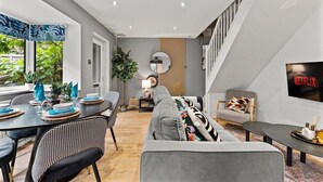 Un salon moderne avec un canapé gris, un coin repas avec une table dressée pour quatre personnes, un escalier et un écran de télévision affichant le logo « NETFLIX ».
