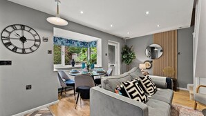 Un salon moderne avec un canapé gris, une grande horloge murale et un coin repas comprenant une table et des chaises.
