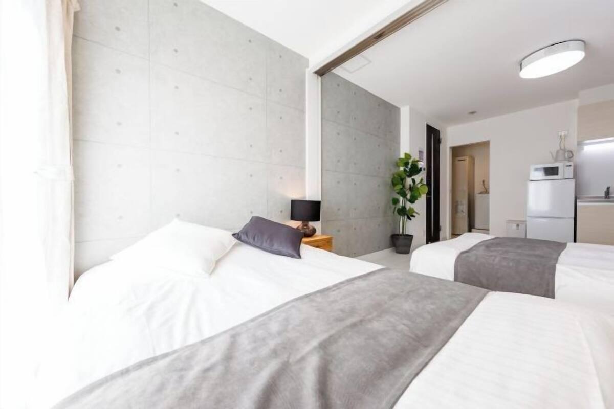 A hotel room with two beds, a bedside table with a lamp, a potted plant, and a kitchenette in the background.