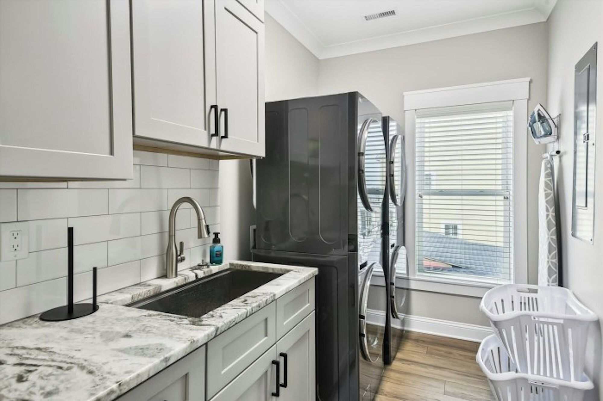 A compact kitchen with white cabinets, a marble countertop, a black appliance, a window with blinds, and a laundry basket.