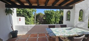 A covered patio with a table and chairs, surrounded by white walls and greenery.