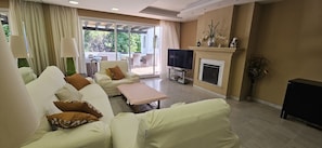 A living room with a sofa set, a flat-screen TV, a fireplace, and a view through a glass door.