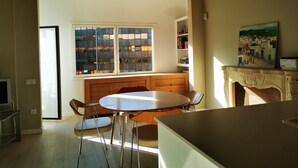 Een eetgedeelte met een ronde tafel en stoelen, een houten bank, een open haard en een raam met uitzicht op gebouwen.