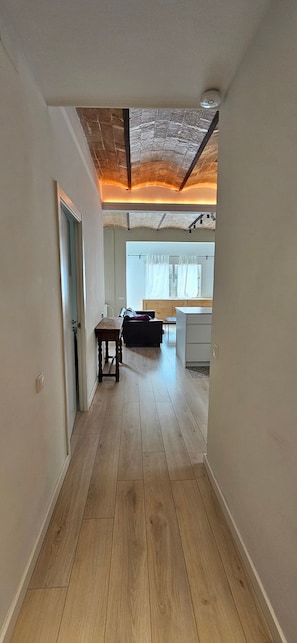 A hallway with wooden flooring leading to a room with a window, a table, and a chair.