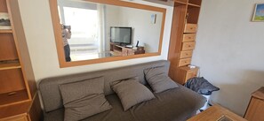 A room with a sofa, a TV on a stand, a mirror, wooden cabinets, and a small table.