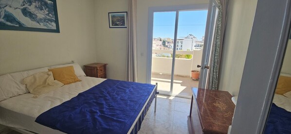 A bedroom with a bed, a small wooden table, a balcony with a view of buildings.