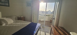 A bedroom with a bed, a wooden nightstand, a window with a view of buildings, and a door leading to a balcony.