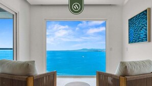 Ein Zimmer mit großem Fenster und Blick auf ein Gewässer, zwei Holzstühle, ein kleiner runder Tisch und ein gerahmtes Kunstwerk an der Wand.