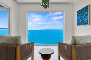 A room with two wooden chairs, a small round table, a large window with a view of the sea, and a framed artwork on the wall.