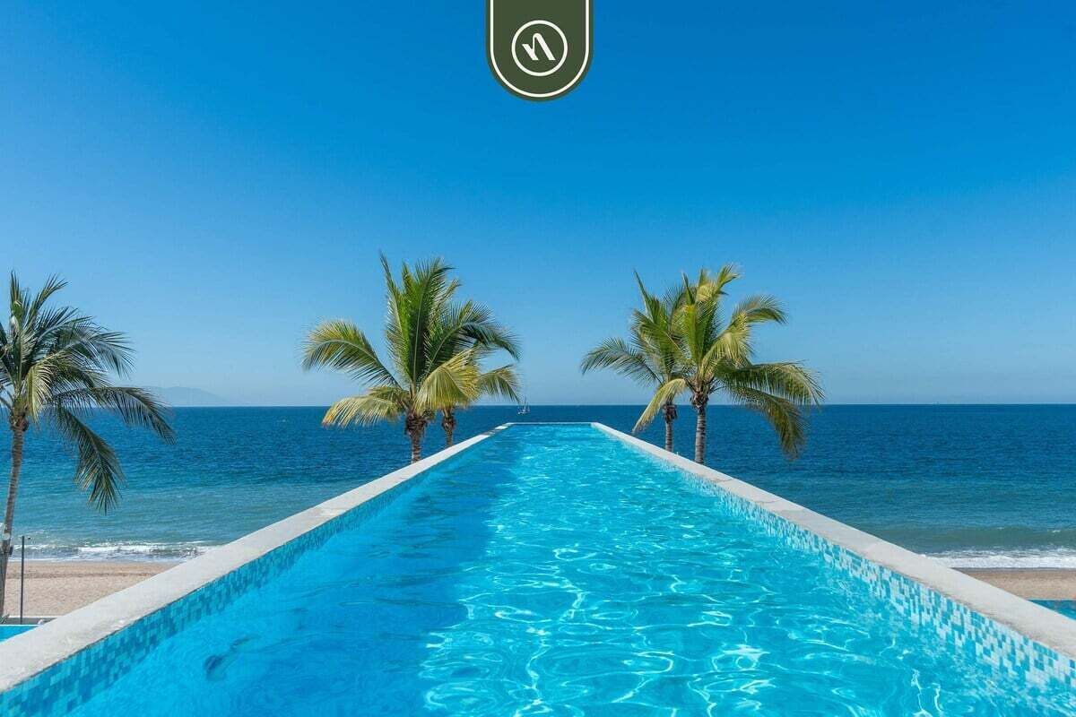 Una piscina larga y estrecha con agua azul cristalina, bordes blancos y que desemboca en una playa con palmeras y el océano al fondo.