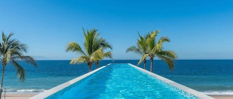 A long, narrow swimming pool with clear blue water, lined with white edges, leading to a beach with palm trees and the ocean in the background.