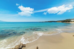 A beach with clear turquoise water, sandy shore, and a distant building on a hill. - Dimora Light House: Elegance and Ocean Views in the Heart of Alghero (Alghero)