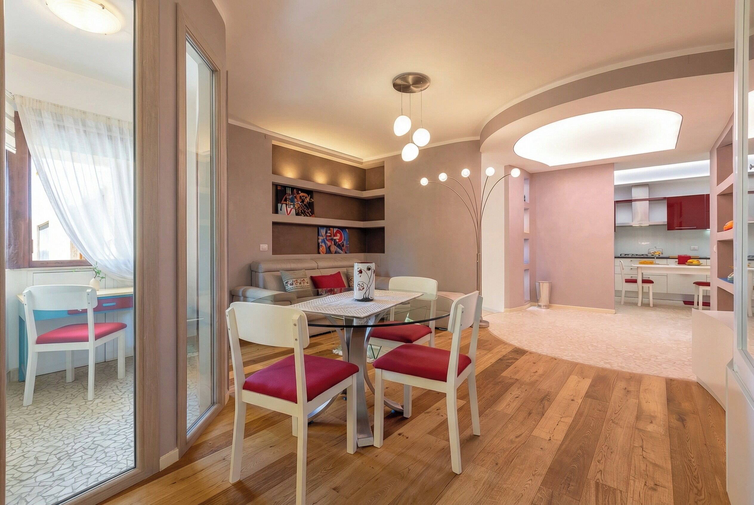 A modern living room with a glass dining table, red cushioned chairs, a sofa, and a kitchen area in the background.