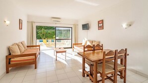 A spacious living area with a wooden sofa, a dining table with chairs, a TV mounted on the wall, and a sliding glass door leading to a balcony with a pool. - EiraSol by Umbral (Albufeira)