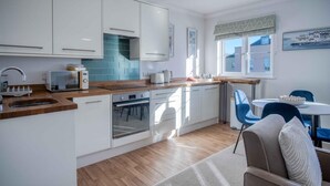 Una cocina moderna con armarios blancos, horno empotrado y microondas. Cuenta con un comedor con mesa y sillas, y un ventanal con vistas a edificios.