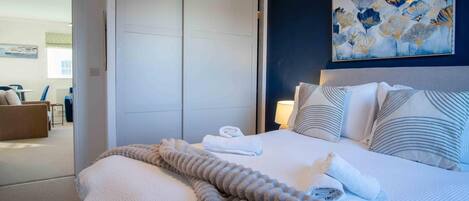 Ett hotellrum med en säng, kuddar, ett täcke, en tavla och utsikt över ett annat rum.