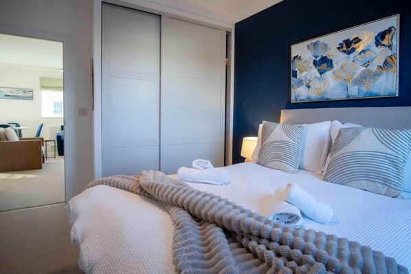 A hotel room with a bed, pillows, a blanket, a painting, and a view of another room. - The Nook North Beach - 1 Bedroom Apartment - Tenby (Tenby)