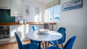 Una cocina moderna con armarios blancos, encimera de madera y una mesa redonda con sillas azules. Hay cuadros en la pared y una ventana con vistas a un pueblo costero.
