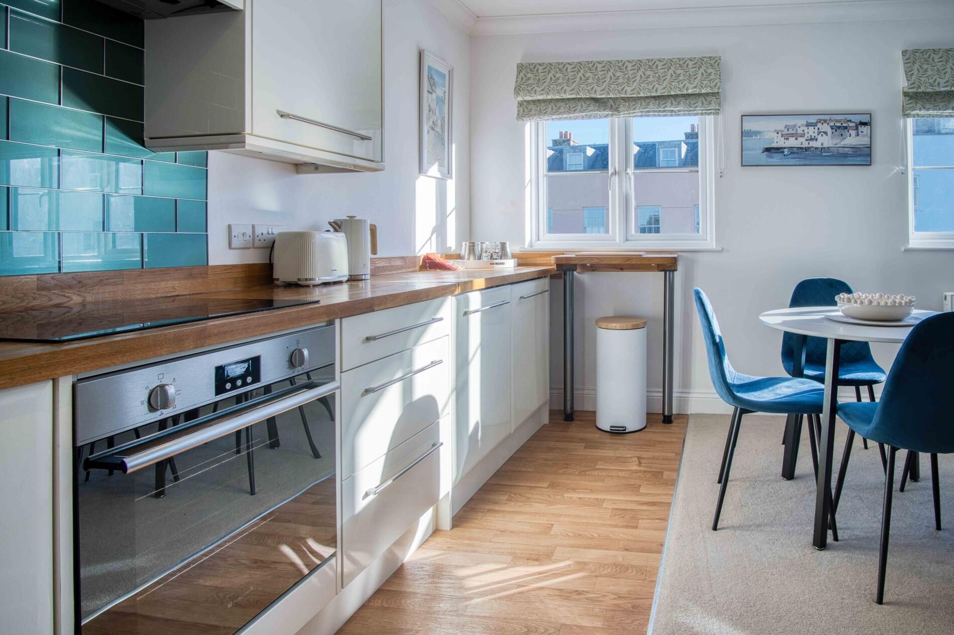 Une cuisine moderne avec des armoires blanches, un comptoir en bois et un four encastré. On y trouve également un coin repas avec une table ronde et des chaises bleues.