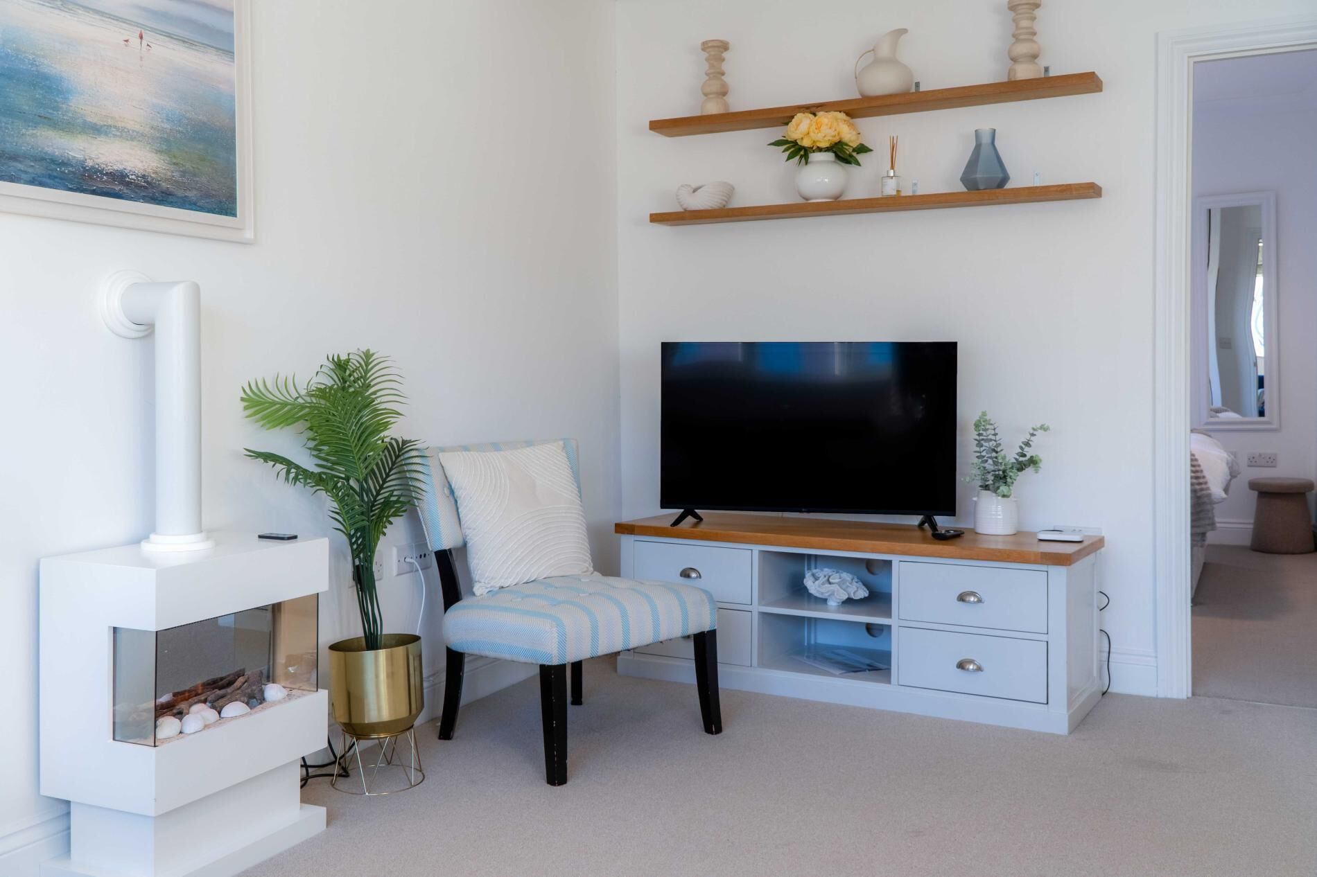 A modern living room with a flat-screen TV, a white fireplace, a striped chair, and a wooden shelf with decorative items.