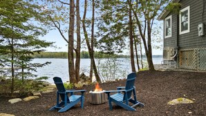Een terras aan het meer met twee Adirondack-stoelen, een vuurkorf en een huis op de achtergrond.