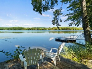 Ein Seeufer mit einer Terrasse, Adirondack-Stühlen und einem Tisch, mit Blick auf einen ruhigen See und bewaldete Hügel.