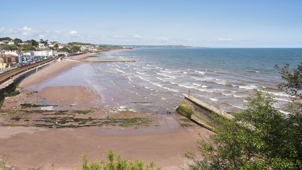 A coastal beach with a promenade, rocky shoreline, and a distant view of a town.
