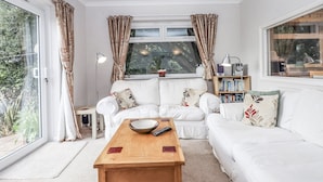 A living room with a white sofa, a wooden coffee table, a bookshelf, and a large window with curtains.