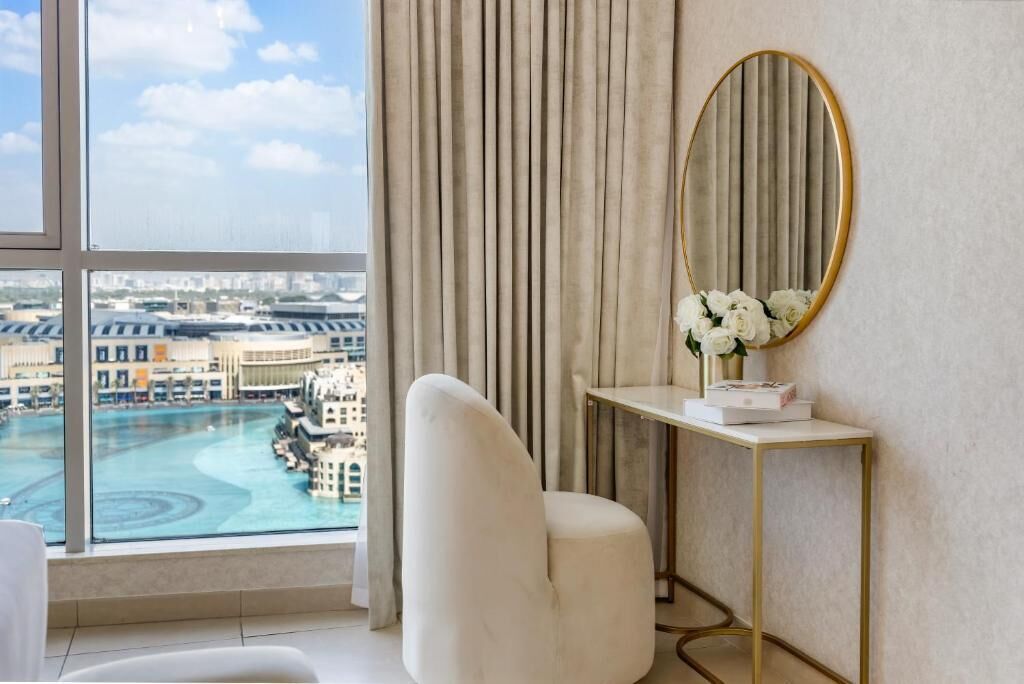 Une chambre d’hôtel moderne dotée d’une grande fenêtre, d’une chaise blanche, d’une console dont le cadre est doré et d’un miroir également doté d’un cadre doré.