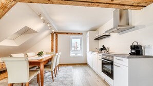 A modern kitchen with white cabinets, a built-in oven, and a coffee machine. There is a dining area with a wooden table and chairs. - Ferienwohnung Alpen Zentral (Bludenz)