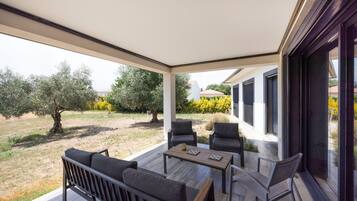 Un patio techado al aire libre con una mesa de madera y dos sillas, con vistas a un jardín con árboles y un edificio al fondo.