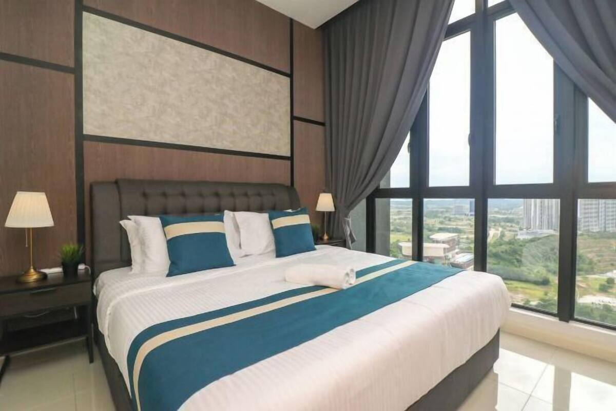 Una habitación de hotel moderna con una cama grande, dos mesitas de noche con lámparas, vistas de la ciudad a través de amplias ventanas y un paisaje urbano visible al exterior.