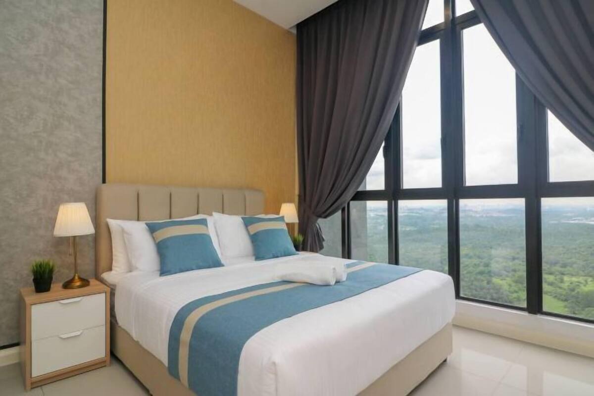 Habitación de hotel con una cama grande, dos mesitas de noche con lámparas y un atril, con vistas a un paisaje a través de amplias ventanas.