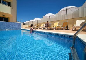 Piscina con suelo azulado de azulejos, tumbonas y sombrillas.