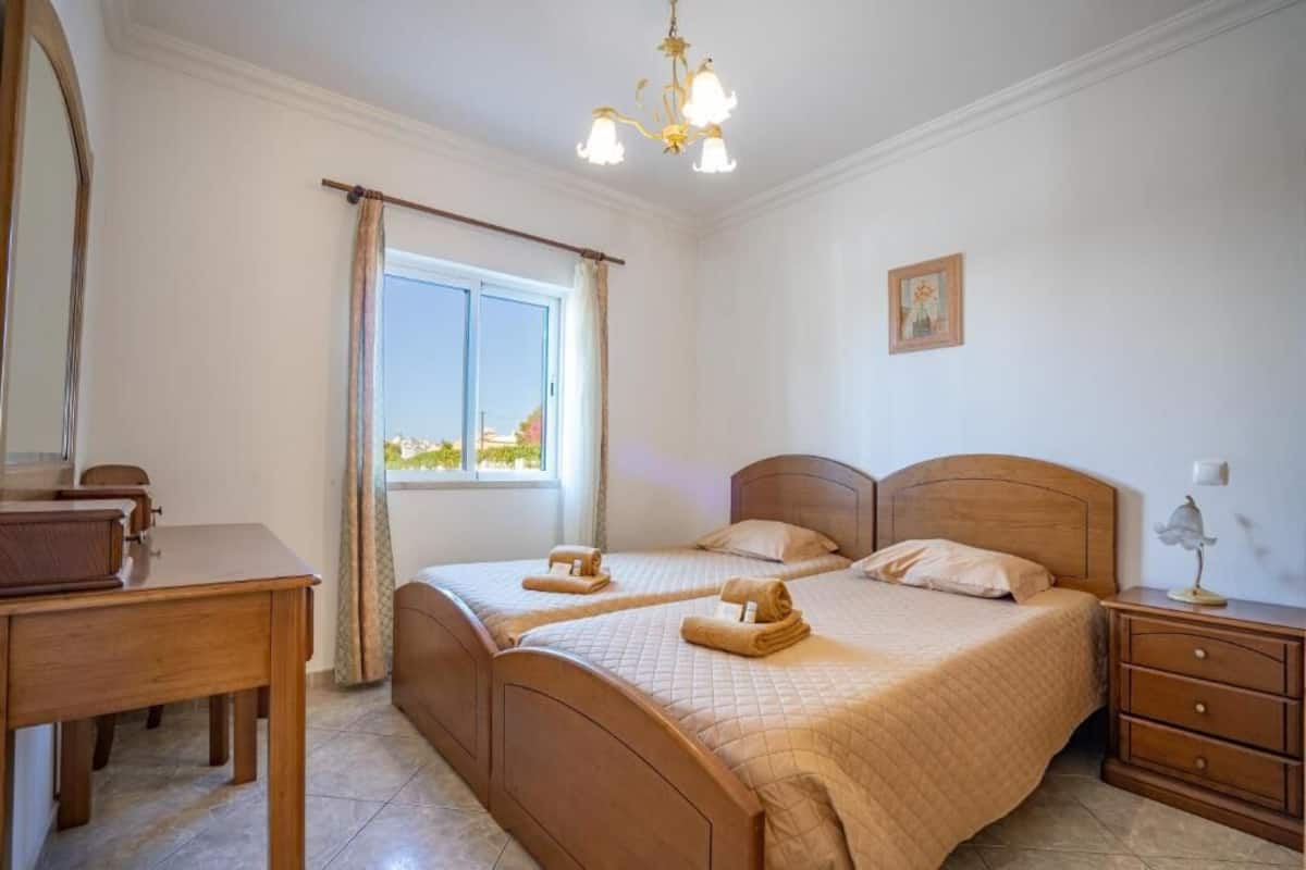 A bedroom with two beds, a desk, a chair, a bedside table, and a window with curtains.