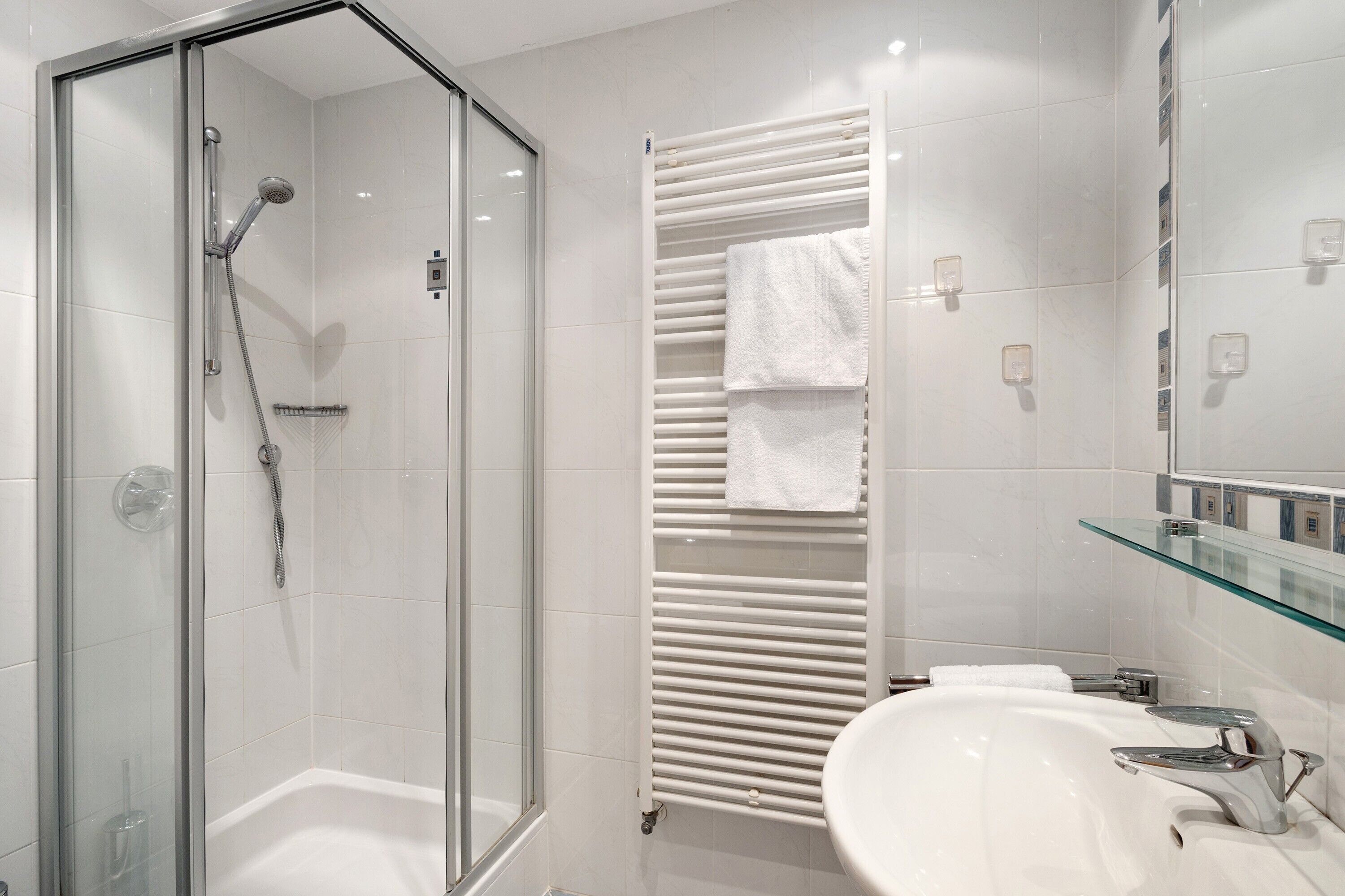 A modern bathroom with a glass-enclosed shower, a white bathtub, a glass-topped vanity, and a towel rack.