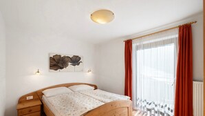 Une chambre à coucher avec un lit en bois, une table de chevet, une lampe fixée au mur, une fenêtre avec des rideaux légers et un radiateur.