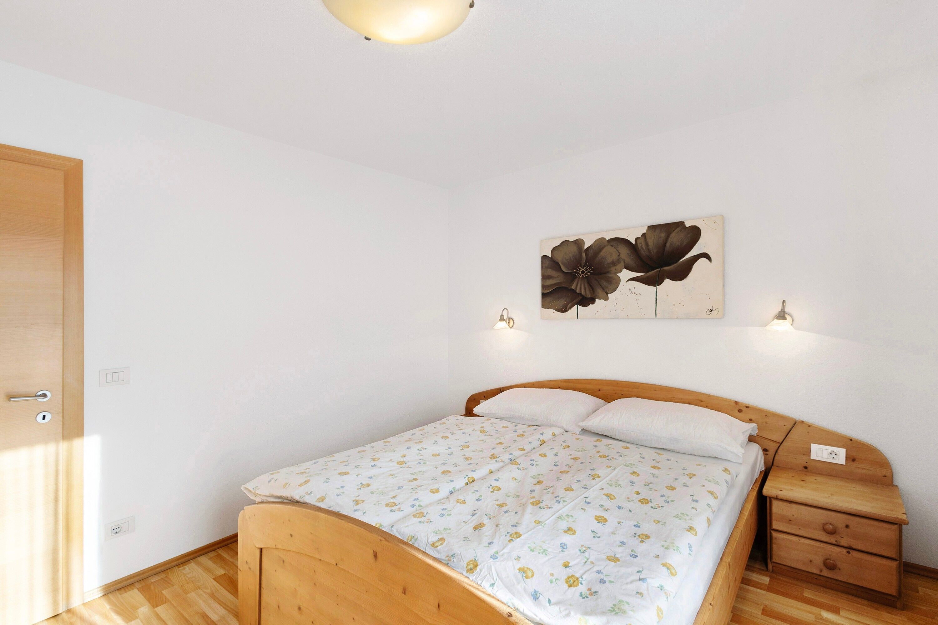 A bedroom with a wooden bed, a nightstand, a wall-mounted lamp, and a floral painting.