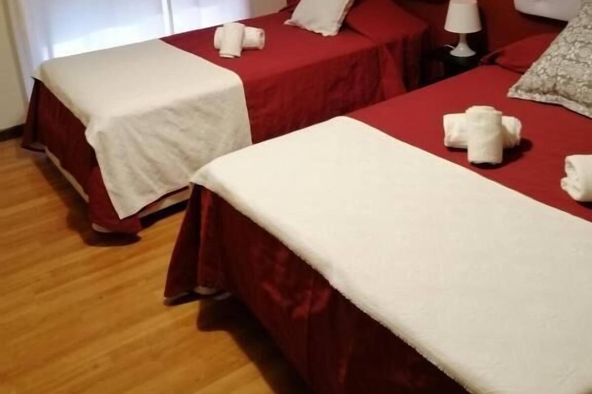 Ένα δωμάτιο ξενοδοχείου με δύο κρεβάτια, το καθένα με λευκό σεντόνι και μαξιλάρια, ένα κομοδίνο με ένα φωτιστικό και ένα παράθυρο με λευκές κουρτίνες.