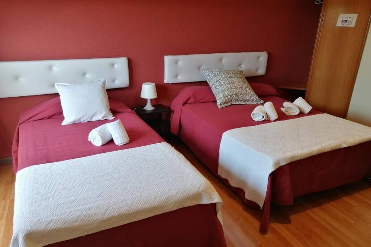 Ένα δωμάτιο ξενοδοχείου με δύο κρεβάτια, το καθένα με λευκό προσκέφαλο, κόκκινα σεντόνια και μια λευκή κουβέρτα. Υπάρχουν δύο μαξιλάρια σε κάθε κρεβάτι, ένα κομοδίνο με ένα φωτιστικό και μια ξύλινη πόρτα με μια επιγραφή.