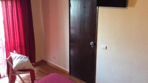 A room with a wooden floor, a dark door, a white chair with a pillow, and a mounted television. - Guest House RSA by Umbral (Loulé)