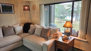 Ein Wohnzimmer mit einem Ecksofa, einem Beistelltisch mit Lampe und einem großen Fenster mit Blick auf einen Garten.