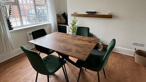 A dining area with a wooden table, four green upholstered chairs, a hanging light fixture, and a shelf above the table.