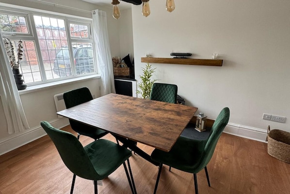 Un coin repas avec une table en bois, quatre chaises à l’assise rembourrée de couleur verte, un luminaire suspendu et une étagère au-dessus de la table.