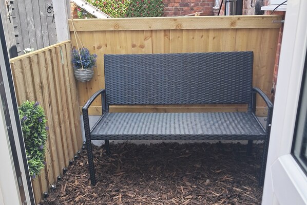 Un banc en métal noir doté d’un dossier en osier, placé dans un espace jardin agrémenté de clôtures en bois et recouvert d’une couche de paillis.