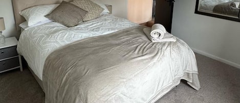 Una cama bien hecha con colcha y almohadas estampadas, una mesita de noche con lámpara y un espejo en la pared.